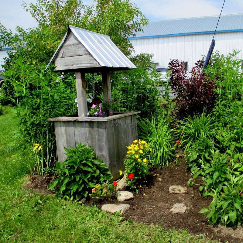 A rustic wooden wishing well stands in a lush garden, surrounded by green plants and colorful flowers, with a blue-roofed building in the background.