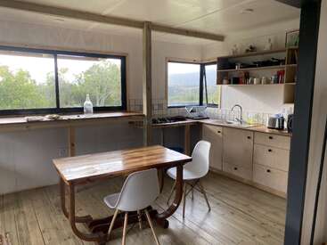 This image shows a cozy, minimalist kitchen with wooden floors, simple cabinetry, a wooden table, two white chairs, open shelves, and large windows overlooking greenery.
