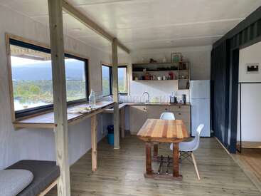 This cozy kitchen has wooden floors, a small dining table with two chairs, lots of natural light from large windows, open shelving, and a bright, minimalist design.