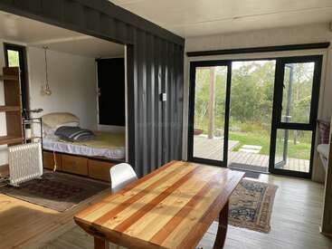 This cozy room features a wooden table, a small bed nook, abundant natural light, sliding glass doors, modern decor, and views of lush greenery outside.