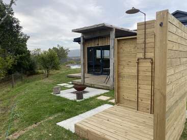 A modern cabin with large windows sits in a grassy yard, featuring a rustic outdoor wooden shower, fire pit, tree stumps for seats, and scenic views.