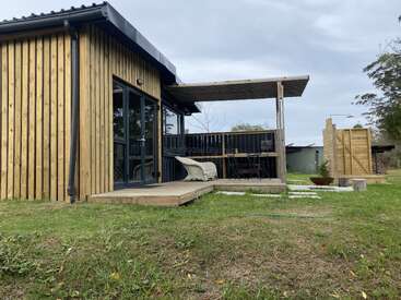 A modern wooden cabin with vertical slats features a small deck, outdoor seating, pergola, grassy yard, fire pit, and rustic outdoor shower area under a cloudy sky.