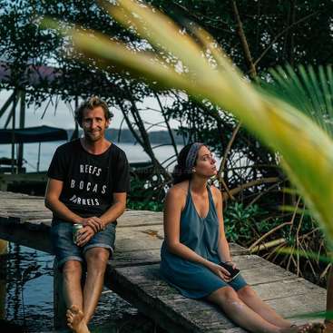 L'image représente un homme et une femme assis sur un quai en bois, entourés d'arbres et d'eau, l'homme tenant une canette et la femme un téléphone.