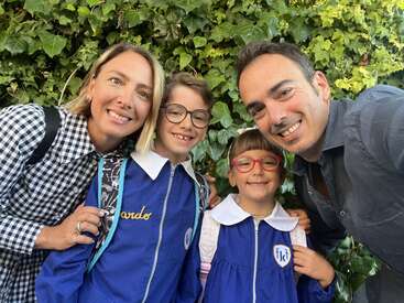 Eine lächelnde Familie, zwei Erwachsene und zwei Kinder in blauen Schuluniformen, posieren gemeinsam im Freien vor einer üppig grünen, mit Efeu bewachsenen Mauer und wirken glücklich.