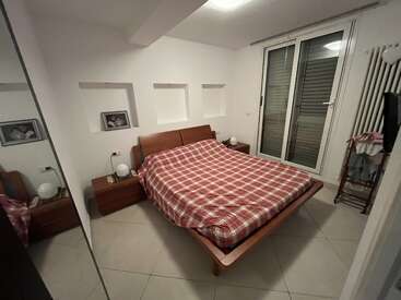 Dieses gemütliche Schlafzimmer verfügt über ein Doppelbett mit einer rotkarierten Decke, Holzmöbel, zwei gerahmte Bilder, große Fenster, Fliesenboden und eine minimalistische Einrichtung.