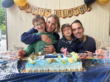Eine fröhliche Familie feiert den 7. Geburtstag eines Kindes mit einer Torte. Dekorationen und Luftballons sorgen für eine festliche Atmosphäre. Alle lächeln und genießen diesen besonderen Moment zusammen.