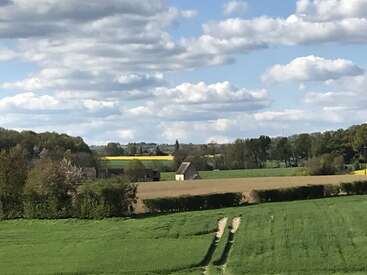 Un paysage champêtre pittoresque, avec des champs verdoyants, des haies, des arbres épars et des bâtiments agricoles, sous un ciel bleu partiellement nuageux, par une journée ensoleillée et lumineuse.