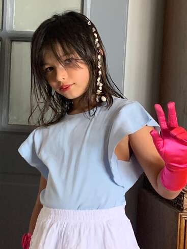 A young girl with dark hair wears a light blue top, white skirt, pink gloves, and beaded hair accessory, flashing a peace sign and smiling slightly.