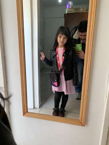 A young girl poses with a peace sign in front of a mirror, dressed in a jacket, dress, and sneakers, while an adult takes the photo.