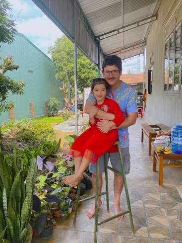 A man and a young girl in a red dress pose happily outdoors. The girl sits on a ladder, surrounded by green plants and sunshine.