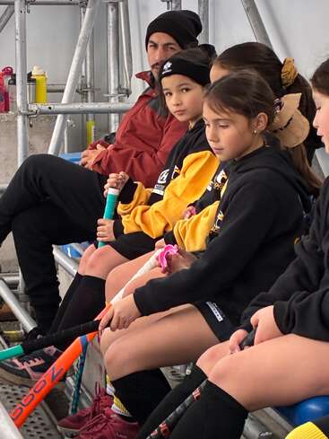 A group of young girls in sports uniforms sit on a bench holding field hockey sticks, accompanied by a coach. They appear focused, waiting for their turn.
