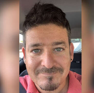 The image is a close-up selfie of a man with short dark hair, wearing a pink polo shirt, in a car.