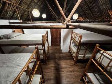 L'image représente une pièce rustique avec un toit de chaume incliné, comprenant quatre lits avec des cadres en bois et des matelas blancs, ainsi qu'une porte en bois à l'extrémité.
