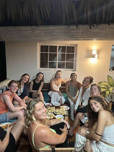 L'image représente un groupe de huit femmes réunies autour d'une table chargée de nourriture, assises sur un canapé et des chaises dans une pièce confortable aux murs blancs et dotée d'une fenêtre.
