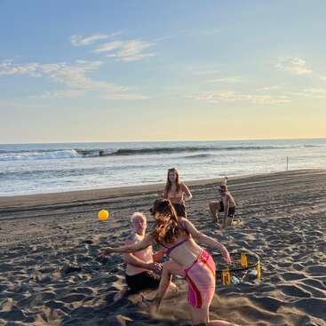Au coucher du soleil, quatre amis jouent au spikeball sur une plage de sable, profitant ensemble des vagues de l'océan, de la chaleur du soleil et d'une ambiance joyeuse et pleine d'énergie.