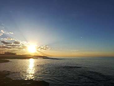 Das Bild zeigt einen heiteren Sonnenuntergang über einem Gewässer, mit einer kleinen Insel oder Halbinsel im Vordergrund und einem blauen Himmel mit Wolken.
