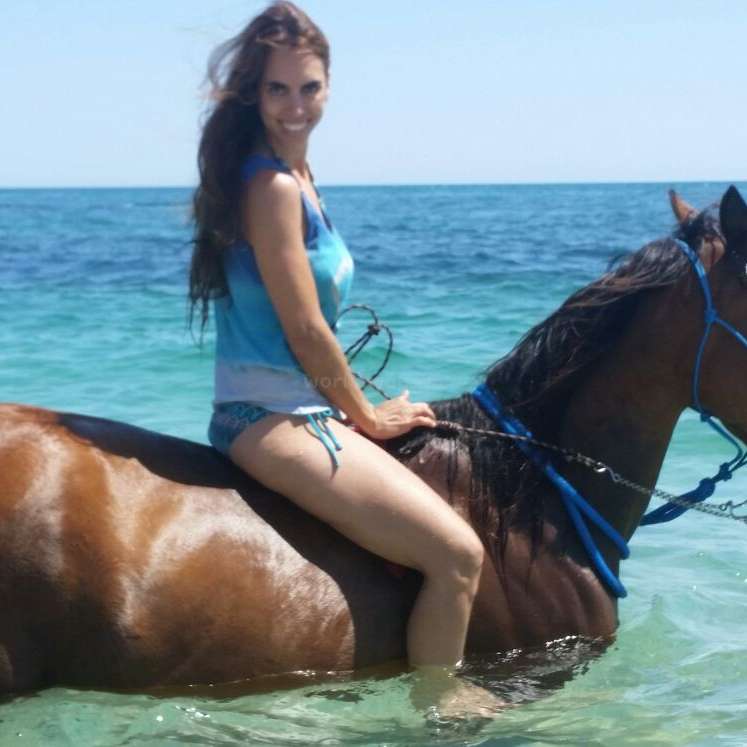 Eine Frau mit langen braunen Haaren sitzt auf einem braunen Pferd im flachen Wasser, schaut nach rechts und trägt ein blaues Tank-Top und eine Bikini-Hose.