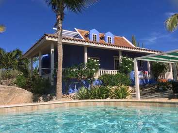 L'image représente une maison bleue sereine avec un toit en tuiles, entourée d'une végétation luxuriante et d'une piscine éclatante, le tout dans un ciel bleu clair.