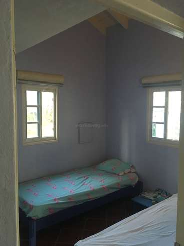 L'image représente une petite pièce avec un lit simple, deux fenêtres, un plafond blanc et un mur bleu avec une subtile teinte violette.