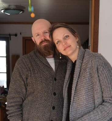 A man and woman stand close together, both wearing gray sweaters. They appear relaxed and content, posing indoors with soft natural light illuminating their faces.