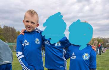 Three young children in matching blue soccer uniforms pose together on a grassy field, smiling. Two faces are digitally obscured; the background is cloudy and green.