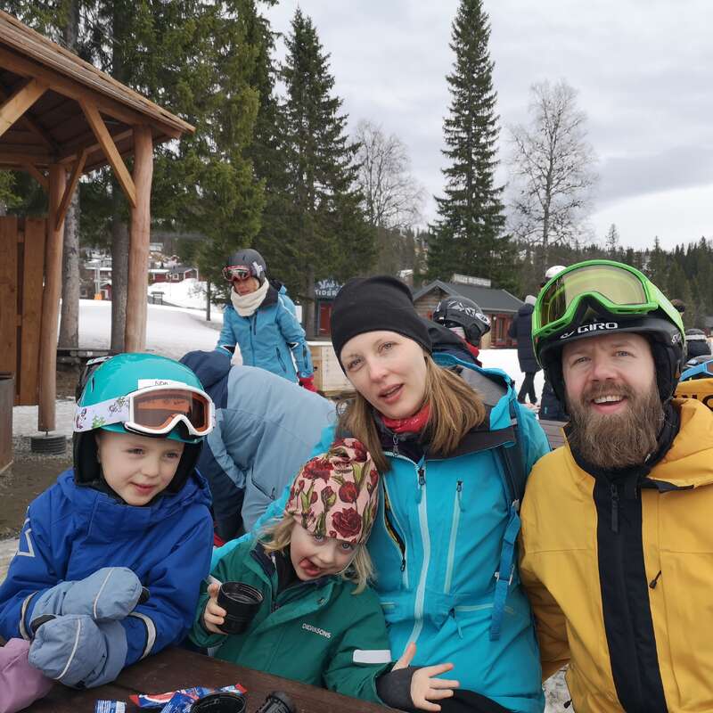 A family of four, dressed in colorful winter gear, sits together outdoors at a ski resort. Snow, trees, and other skiers can be seen in the background.