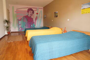 Ein Zimmer mit drei Einzelbetten, bunten Tagesdecken, Holzboden, einem Wandbild, auf dem eine Person eine Blume hält, einer Topfpflanze und gerahmten Landschaftsfotos.