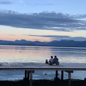 The image depicts a serene scene of a man and child sitting on a wooden dock, gazing out at a tranquil lake and majestic mountains in the distance.