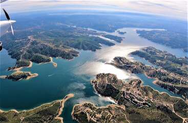 Esta foto aérea de tirar o fôlego mostra um lago panorâmico com margens sinuosas, colinas verdejantes, penínsulas espalhadas, povoados distantes e a luz do sol brilhando na água azul tranquila.