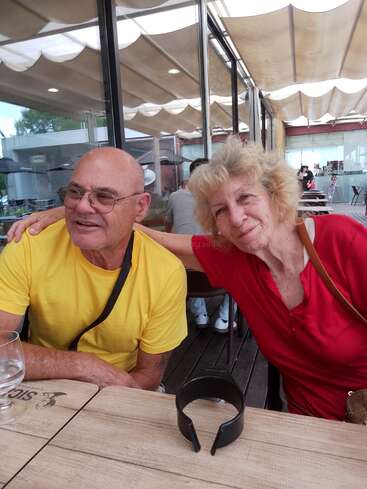 Um casal de idosos senta-se junto em um café ao ar livre. O homem usa uma camisa amarela e a mulher, vermelha. Eles sorriem, demonstrando cordialidade e felicidade, aproveitando seu tempo.