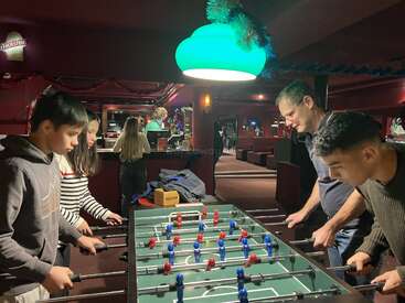 Quatre personnes jouent au baby-foot dans une pièce confortable, chaleureusement éclairée et décorée dans des tons marron. D'autres personnes sont visibles à l'arrière-plan, et des décorations festives sont suspendues au-dessus.