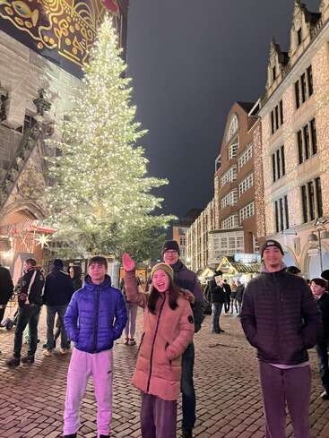 Quatre personnes posent joyeusement devant un grand sapin de Noël illuminé dans une rue festive de la ville, remplie de lumières, de bâtiments historiques et de décorations de Noël.