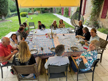 A group of people sits around a large, rectangular table covered with a colorful tablecloth, enjoying a meal together outdoors in a lush garden setting.
