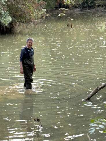 Un homme se tient debout, tout sourire, dans une eau peu profonde et trouble, entouré d'arbres. Vêtu de cuissardes imperméables et d'un sweat à capuche, il fait des vagues tout en profitant du cadre naturel.