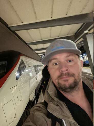 Um cara de boné cinza e jaqueta marrom tira uma selfie na plataforma da estação, ao lado de um trem moderno de alta velocidade branco e vermelho.
