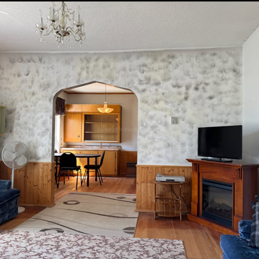 Sala de estar aconchegante com móveis azuis, dois tapetes, abajur vintage, TV sobre a lareira, ventilador, lustre e arco que leva à área de jantar retrô e à cozinha.