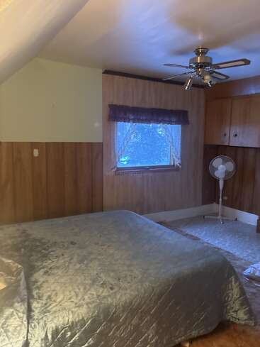 Esse quarto aconchegante tem paredes com painéis de madeira, um ventilador de teto prateado, uma janela com cortinas, um ventilador de canto e uma cama coberta com roupa de cama prateada.