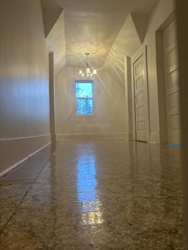 Um corredor elegante com piso brilhante, paredes creme e várias portas. Uma janela no final deixa entrar a luz natural, iluminada por um lustre acima.