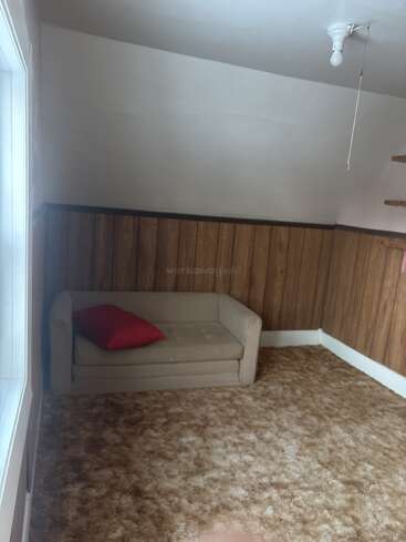Esse quarto tem paredes com painéis de madeira, carpete bege, um sofá pequeno bege com uma almofada vermelha, uma lâmpada exposta e uma janela que deixa a luz entrar.