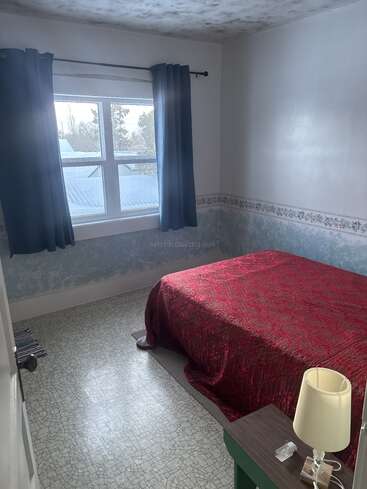 Um quarto pequeno e aconchegante com uma colcha vermelha, mesa lateral, abajur e uma janela com cortinas azuis que deixam a luz do sol entrar. O piso tem um padrão de azulejos.