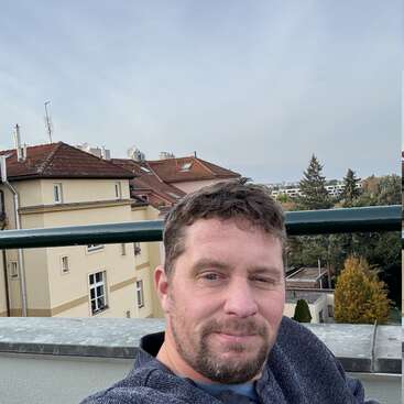 Ein Typ mit kurzen braunen Haaren und Bart macht ein Selfie auf einem Balkon. Hinter ihm sieht man Gebäude mit roten Dächern und Bäume unter einem bewölkten Himmel.