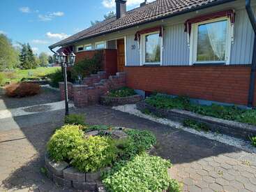 The image depicts a charming one-story house with a brown tiled roof, white walls, and red brick accents, surrounded by a beautifully landscaped yard.