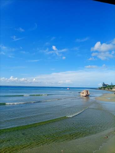 Das Bild zeigt eine ruhige Strandszene mit einem kleinen Boot am Ufer, vor dem Hintergrund eines klaren blauen Himmels mit vereinzelten Wolken und einer ruhigen See.