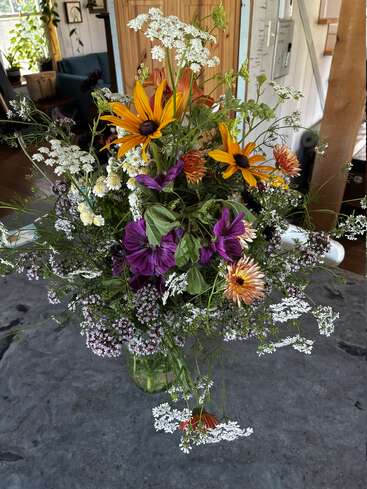 Um buquê vibrante de flores silvestres em um jarro de vidro repousa sobre uma superfície de pedra cinza, com flores amarelas, roxas, laranjas e brancas, dentro de uma casa aconchegante.