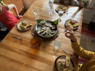 Quatro pessoas estão sentadas ao redor de uma mesa de madeira, com pratos de frutos do mar, pão de alho e copos de água. No centro, há uma grande travessa de ostras.