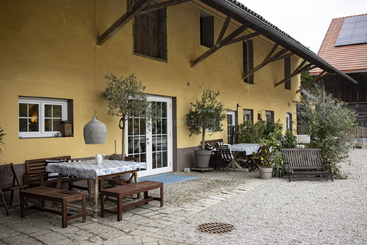 Um charmoso pátio ao ar livre tem mesas de madeira, bancos, vasos de plantas e toalhas de mesa ao lado de um prédio amarelo rústico, convidando você a relaxar num ambiente tranquilo no campo.