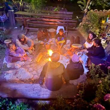Un groupe de personnes est assis en cercle autour d'un feu de camp, la nuit, en pleine nature, entouré de tambours, de couvertures et d'une ambiance chaleureuse et joyeuse en plein air.
