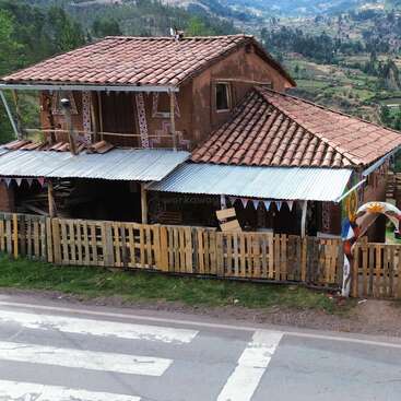 A rustic house with a tiled roof and wooden fence sits along a road in a hilly rural area, decorated with colorful pennant banners and artwork.