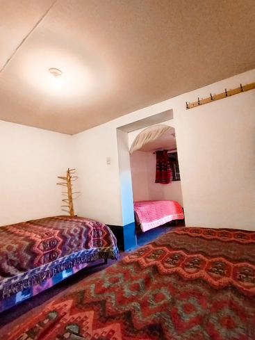 Cette chambre confortable comprend deux lits aux motifs colorés au premier plan, un porte-manteau en bois, des murs blancs et une petite pièce attenante dans laquelle on aperçoit un autre lit.