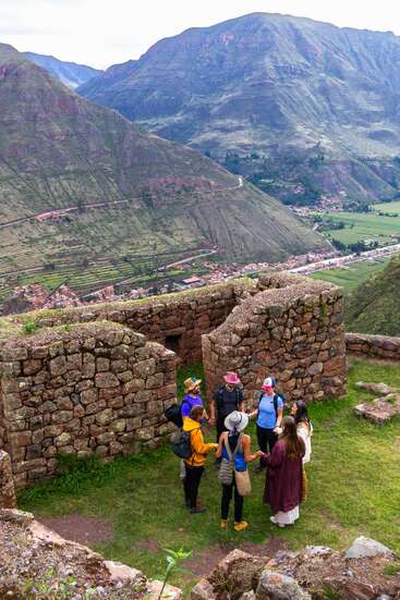 Un groupe de personnes se tient en cercle parmi les ruines de pierres anciennes sur une colline herbeuse, entourée de montagnes verdoyantes et d'une vallée pittoresque.